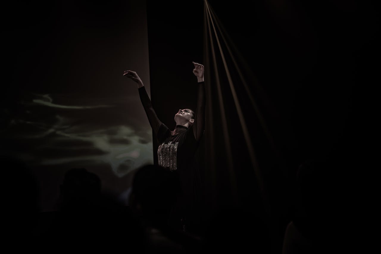 about-us-01 A ballerina elegantly stretches on stage, illuminated by a spotlight, showcasing the art of ballet.